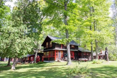 A rustic wooden house with red accents surrounded by lush green trees and a grassy lawn.