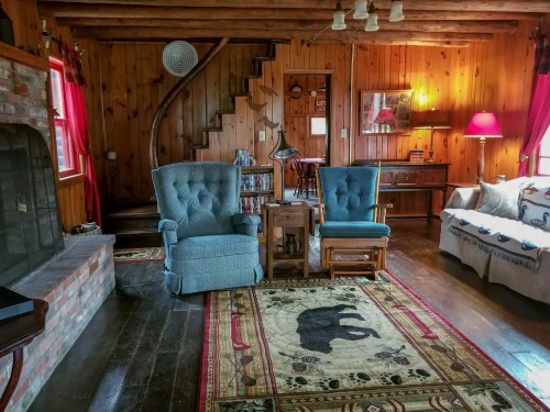 Cozy wooden living room with two blue chairs, a rug featuring a bear, and a spiral staircase in the background.