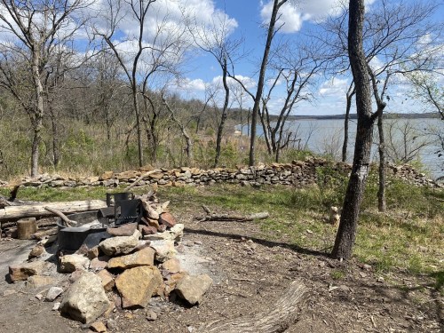A serene lakeside campsite with a stone fire pit, surrounded by trees and a view of the water under a blue sky.