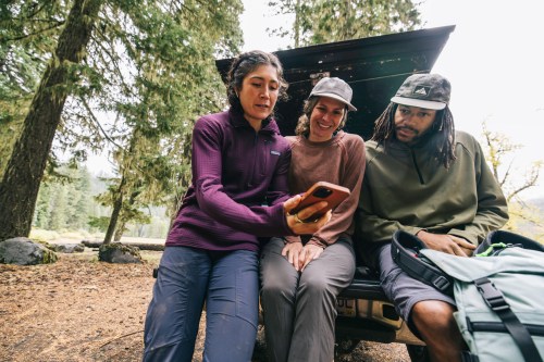Three friends sitting on a truck tailgate in a forest, looking at a phone together, surrounded by trees.