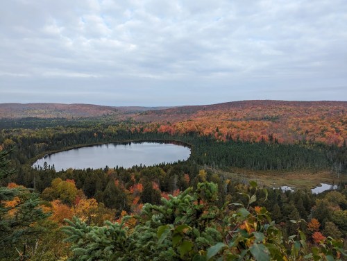 A scenic view of a circular lake surrounded by vibrant autumn foliage and rolling hills under a cloudy sky.