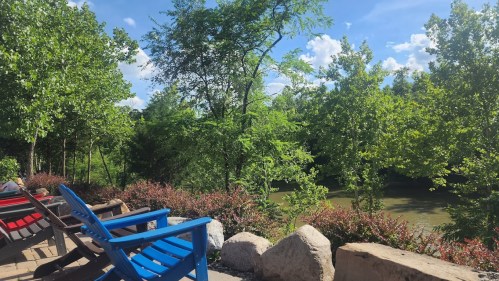 A serene riverside view with blue and red chairs, lush green trees, and a clear blue sky.
