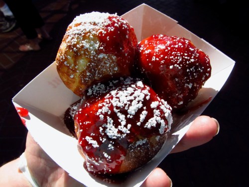 A hand holding a tray of three fried dough balls topped with powdered sugar and red fruit sauce.