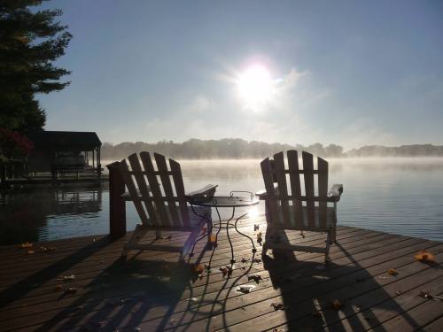 Two wooden chairs on a dock by a misty lake, with the sun rising in the background and autumn leaves scattered on the wood.