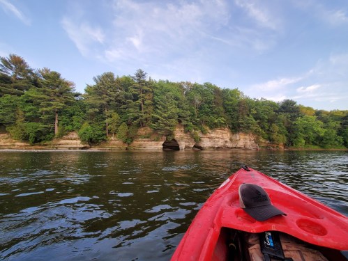 A red kayak floats on calm water, with lush green trees and a rocky cliff in the background under a blue sky.