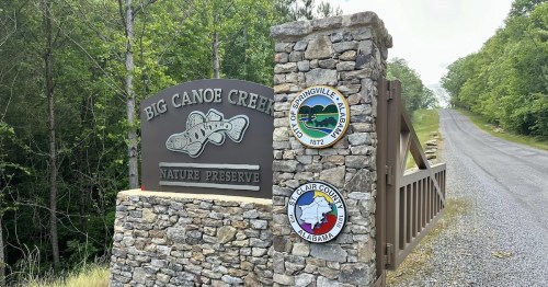 Sign for Big Canoe Creek Nature Preserve, featuring stone structure and local government seals, with a gravel road ahead.