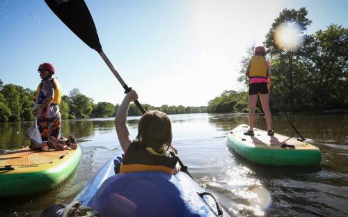 A group of people kayaking on a sunny day, with one child paddling and another standing on a paddleboard.