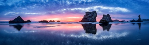 A panoramic view of a beach at sunset, featuring rock formations and colorful reflections in the wet sand.