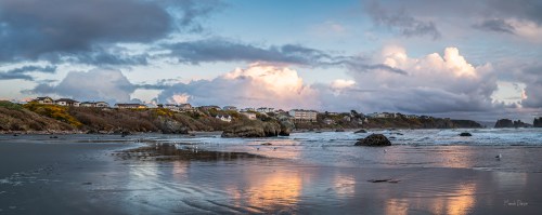 A panoramic view of a beach with rocky formations, reflecting clouds and houses on a coastal cliff under a colorful sky.