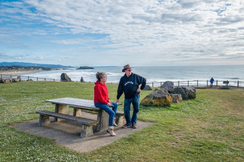 A man and woman stand by a picnic table overlooking a scenic beach with waves and clouds in the background.