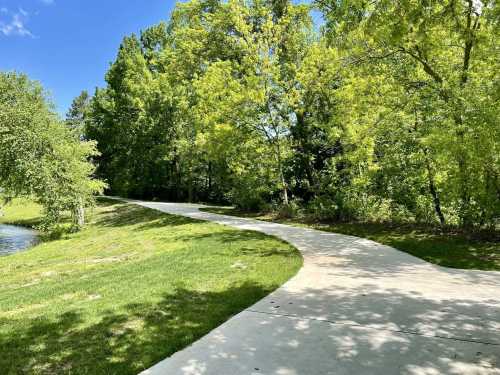 A winding concrete path through lush green trees beside a calm waterway under a clear blue sky.