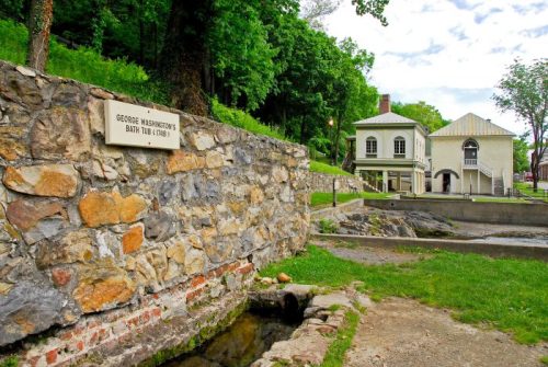 Stone structure with a plaque reading "George Washington's Bath Tub (1783)" near a historic building and greenery.
