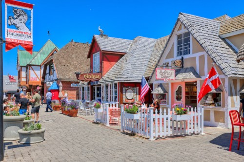 Colorful storefronts line a sunny street, with people enjoying the atmosphere and flags displayed outside.