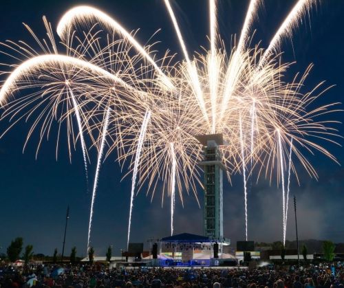 Fireworks illuminate the night sky above a concert stage, with a large crowd gathered below.