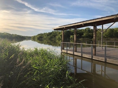 A wooden dock extends over a calm river, surrounded by lush greenery and a serene sky.