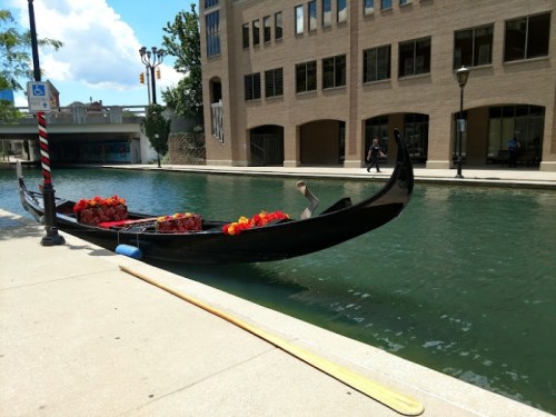 A gondola with flowers is moored by a canal, with buildings and a person walking in the background.