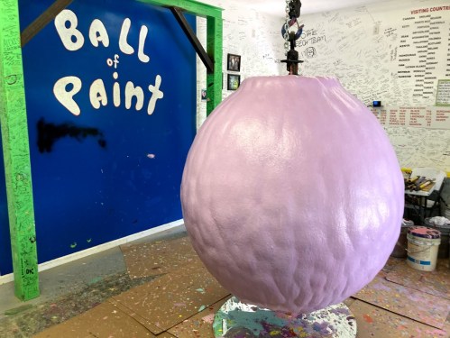 A large pink ball of paint hangs in a colorful room with graffiti-covered walls and a blue backdrop reading "BALL of PAINT."