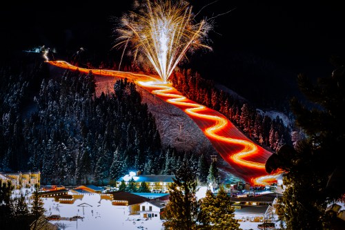 A snowy mountain slope illuminated by fireworks, with glowing trails from skiers winding down the hill at night.