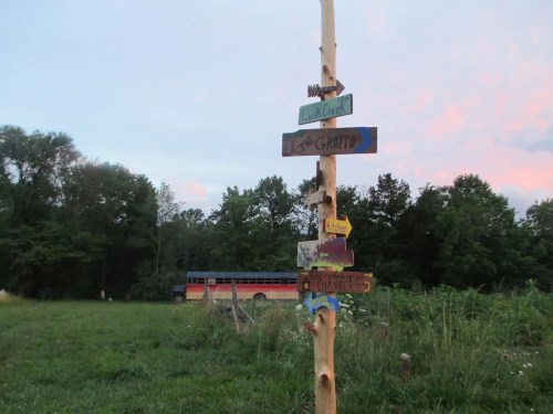 A wooden signpost with colorful directional signs in a grassy area, with a bus and trees in the background.
