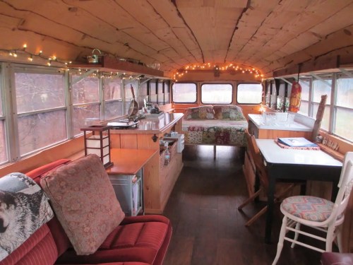 Cozy interior of a converted bus, featuring a bed, seating area, and warm string lights.
