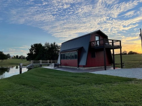 A red and gray house by a pond, with a deck, surrounded by green grass and a sunset sky filled with clouds.