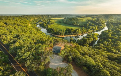 Aerial view of a winding river surrounded by lush greenery and rolling hills, with a road visible in the foreground.