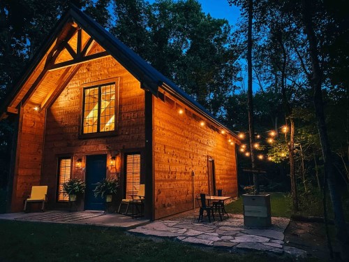 A cozy wooden cabin illuminated at night, surrounded by trees and string lights, with a welcoming porch and outdoor seating.