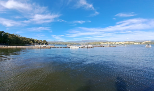 A serene lake scene with boats docked, surrounded by green hills and a clear blue sky with scattered clouds.