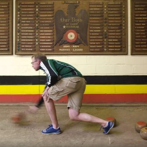A person in a green jacket and shorts rolls a bowling ball on a lane, with a wall of plaques in the background.