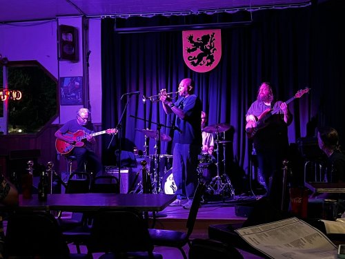 A live band performs on stage, featuring a trumpeter, guitarist, bassist, and drummer, with a purple-lit backdrop.