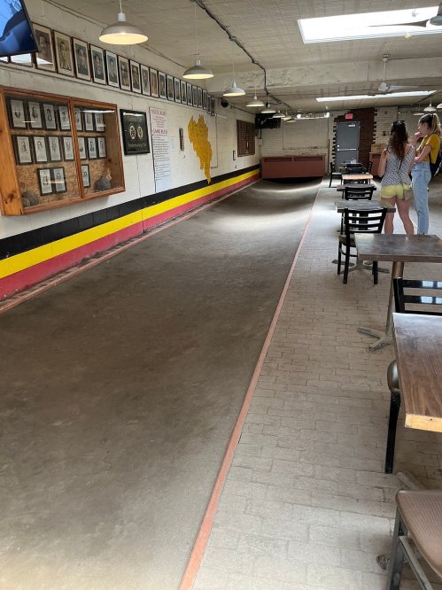 A spacious indoor bowling alley with a smooth lane, framed photos on the wall, and two people standing nearby.