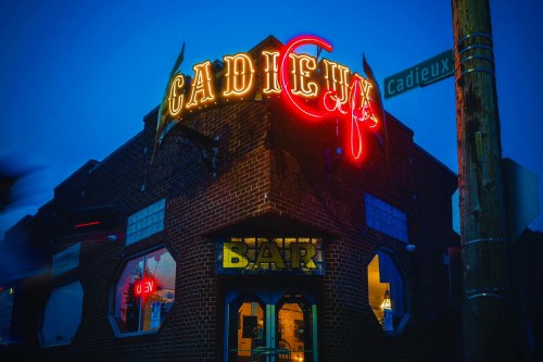 Neon sign of Cadieux Bar on a brick building at dusk, with a street sign visible nearby.
