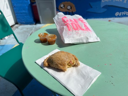 A green table with a stuffed pastry on a napkin, two sauce cups, and a takeout bag in a colorful outdoor setting.