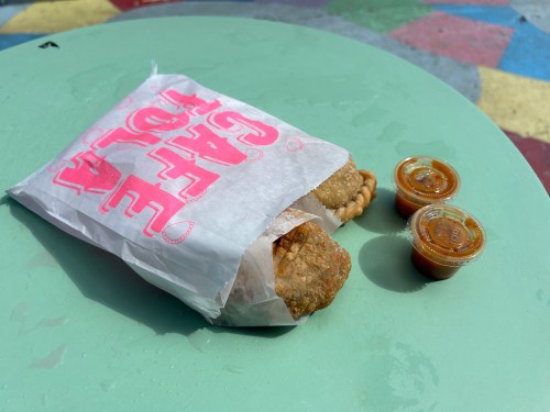 A wrapped fried pastry on a green table, accompanied by two small containers of sauce.