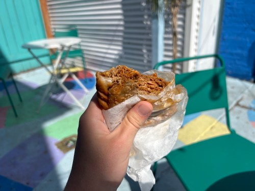 A hand holding a sandwich wrapped in paper, with a colorful outdoor setting in the background.