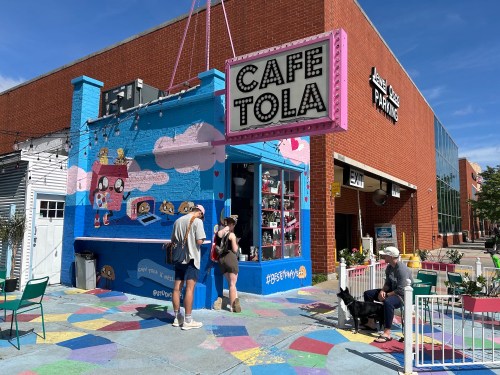 A colorful café exterior with a vibrant mural, people walking by, and a dog on a sunny day.