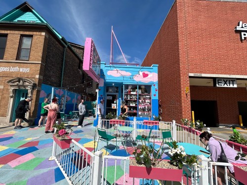 A colorful storefront with a mural, surrounded by people and outdoor seating, under a clear blue sky.