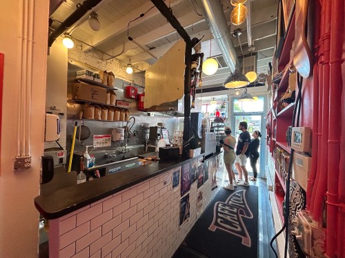 A cozy café interior with a counter, kitchen area, and two customers chatting near the entrance. Bright and inviting atmosphere.