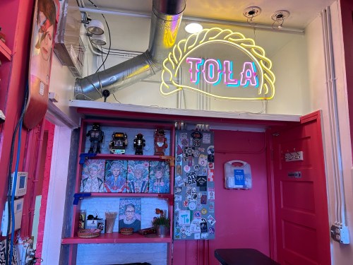 A colorful interior of a cafe featuring a neon sign that reads "TOLA," with eclectic decor and shelves of art.