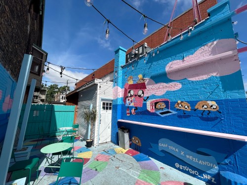Colorful outdoor café space with mural art, string lights, and green tables and chairs under a blue sky.