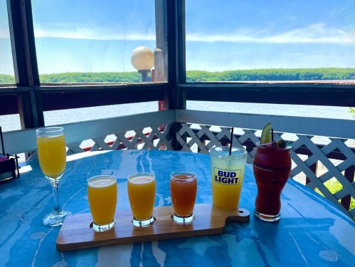 A table with colorful drinks, including mimosas, a cocktail, and a beer, with a scenic view of water and greenery.