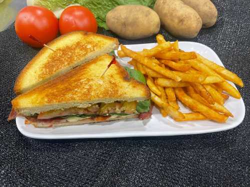 A toasted sandwich with lettuce, tomato, and bacon, served with crispy French fries and fresh vegetables in the background.