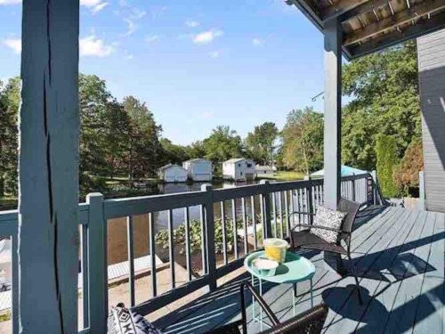 A serene lakeside view from a porch with chairs and a small table, surrounded by trees and houses.