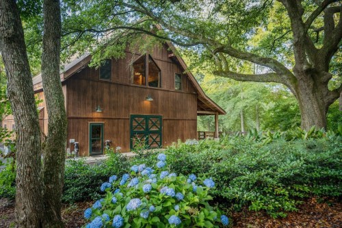 A rustic wooden barn surrounded by lush greenery and blue hydrangeas under a canopy of trees.