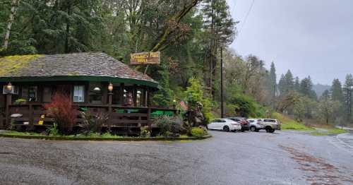 A cozy cabin-style grill surrounded by lush greenery on a rainy day, with parked cars nearby.