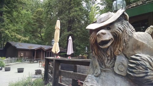 A wooden bear statue wearing a hat stands near a rustic outdoor area with umbrellas and cabins in a forest setting.