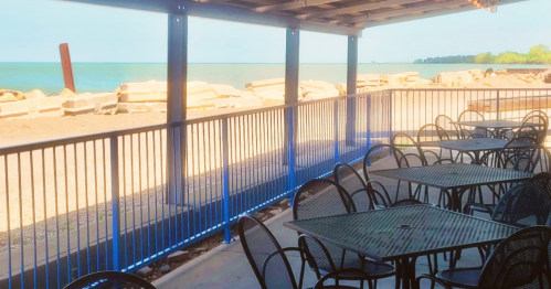 Outdoor seating area with black tables and chairs overlooking a lake and rocky shoreline under a covered patio.