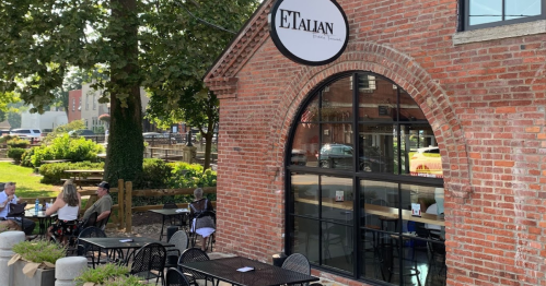 Exterior of a brick restaurant with a sign reading "ETALIAN," featuring outdoor seating and a green park area nearby.