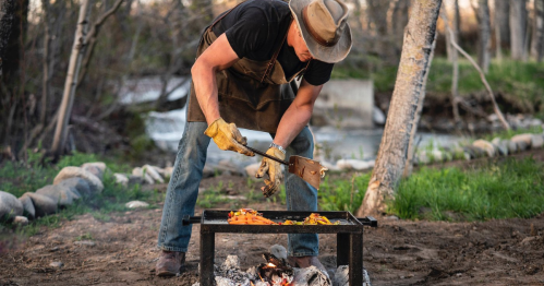 A person in a hat and apron grilling food over an open fire in a wooded area by a stream.