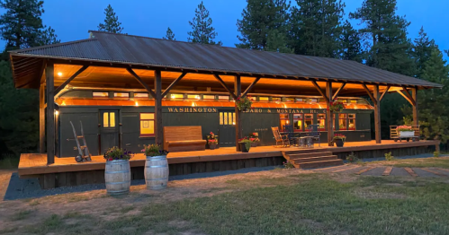 A rustic building with warm lights, surrounded by trees, featuring a porch and barrels, set in a serene evening landscape.